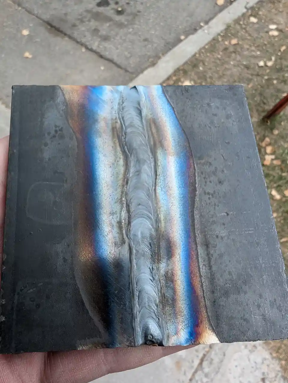 A hand holds a dark metal plate with a central weld seam showing iridescent blue, orange, and purple heat-affected zones…