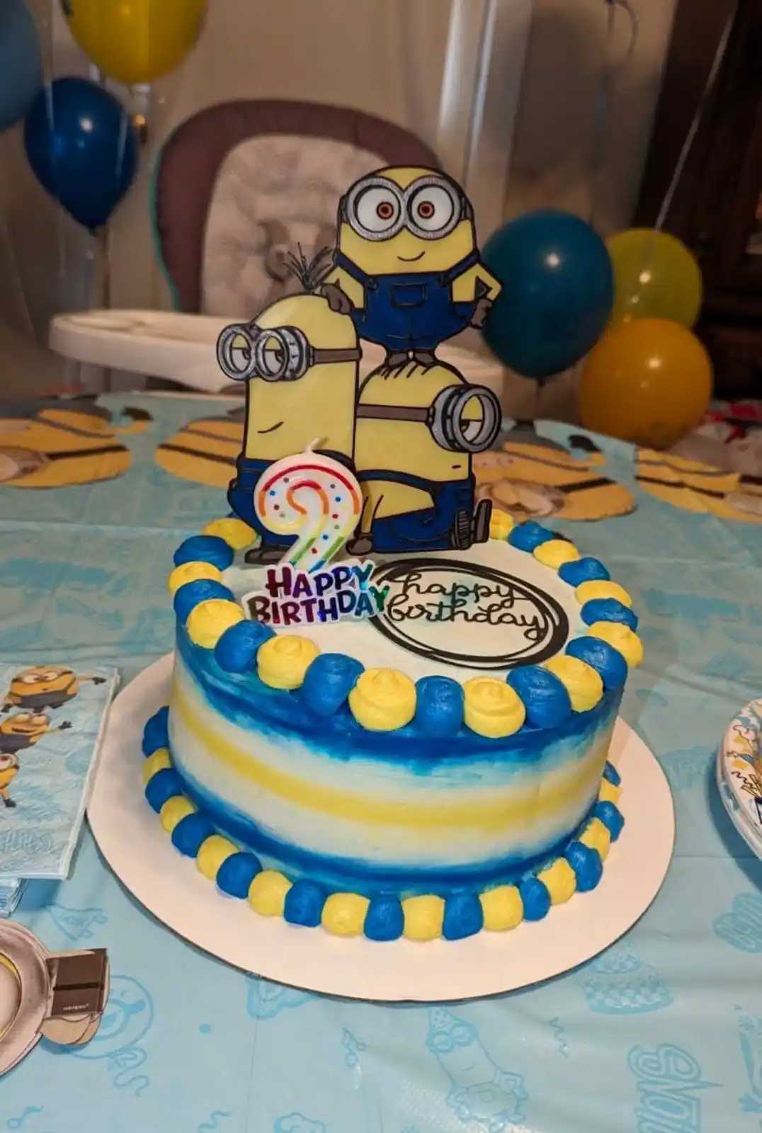 A birthday cake decorated with blue and yellow frosting, topped with Minion figurines and a candle shaped like the number 2…