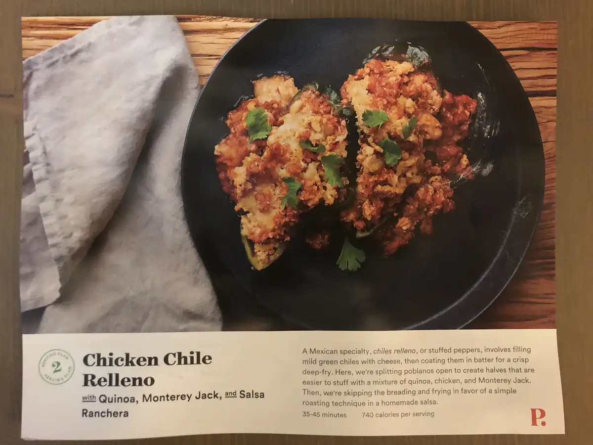 A printed recipe card shows a dish of chicken chile relleno on a black plate, garnished with cilantro, next to a gray cloth…