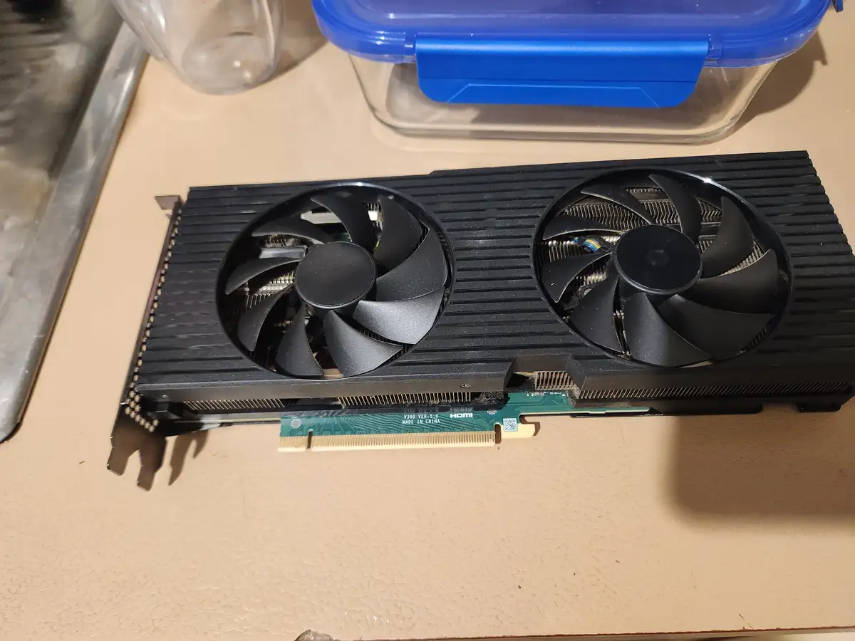 A black dual-fan graphics card with a green circuit board and gold PCIe connector lies on a beige countertop, with a…