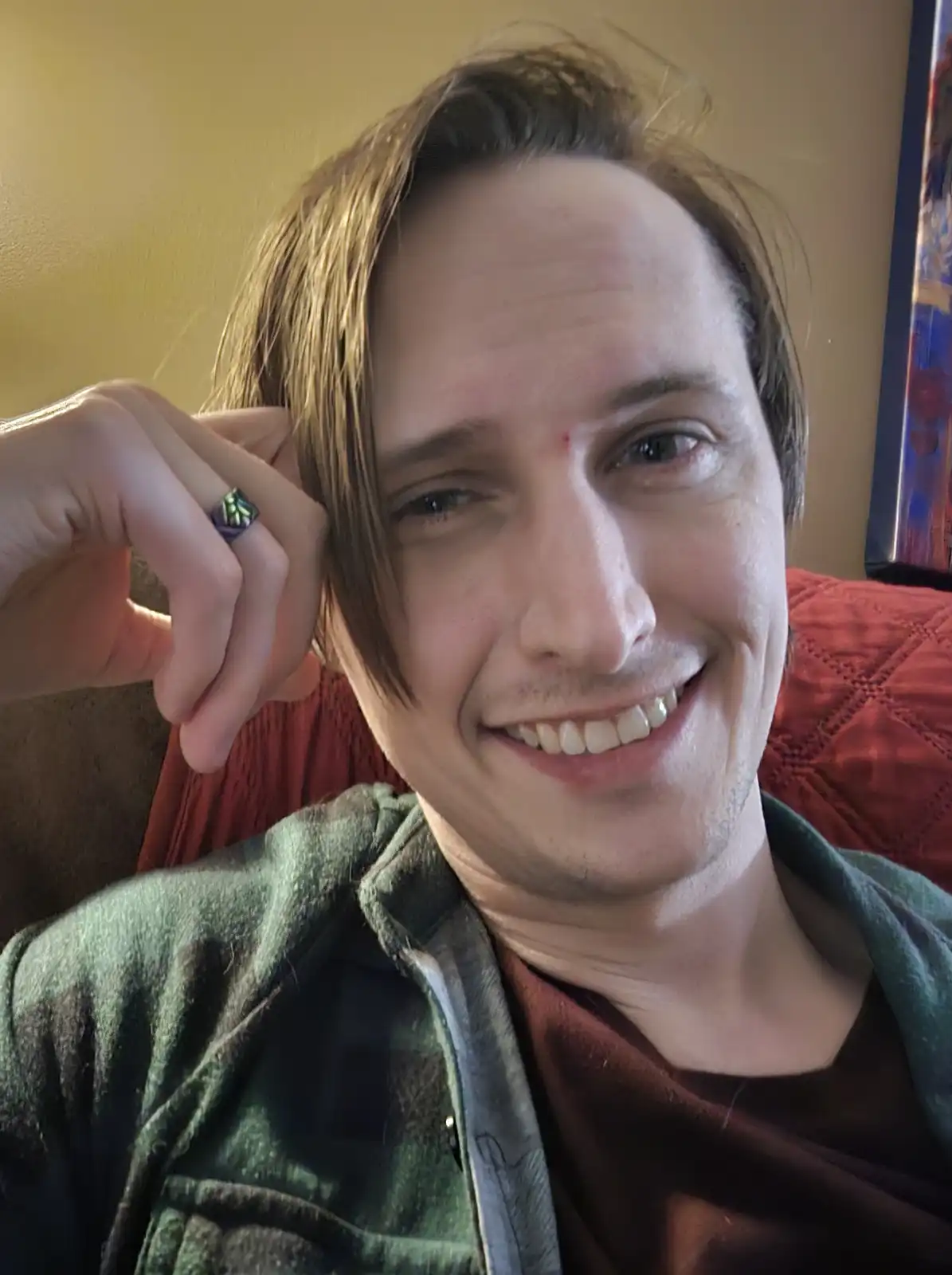 A smiling man with medium-length brown hair, wearing a green plaid shirt over a red shirt, holds up his left hand to show a…