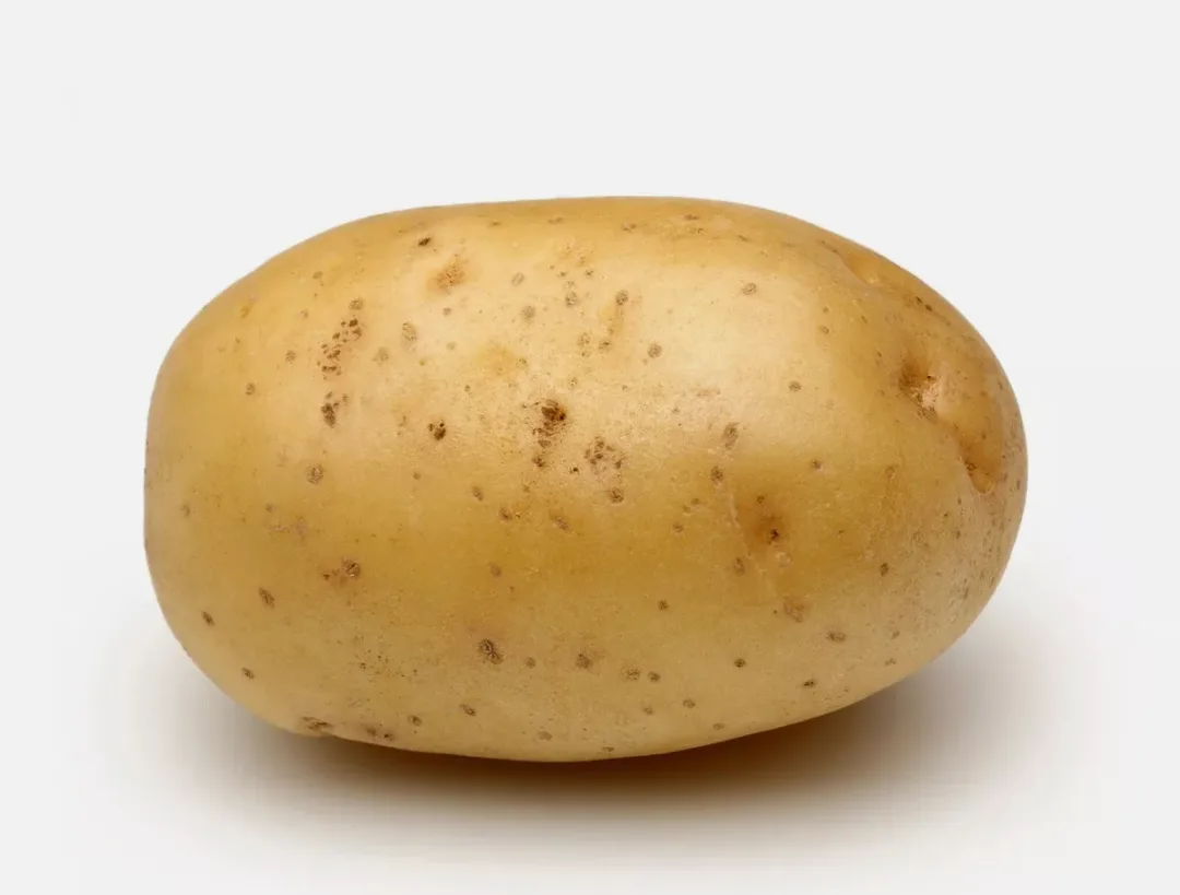 A single, unpeeled, light brown potato with visible eyes and blemishes sits centered against a plain white background. The…