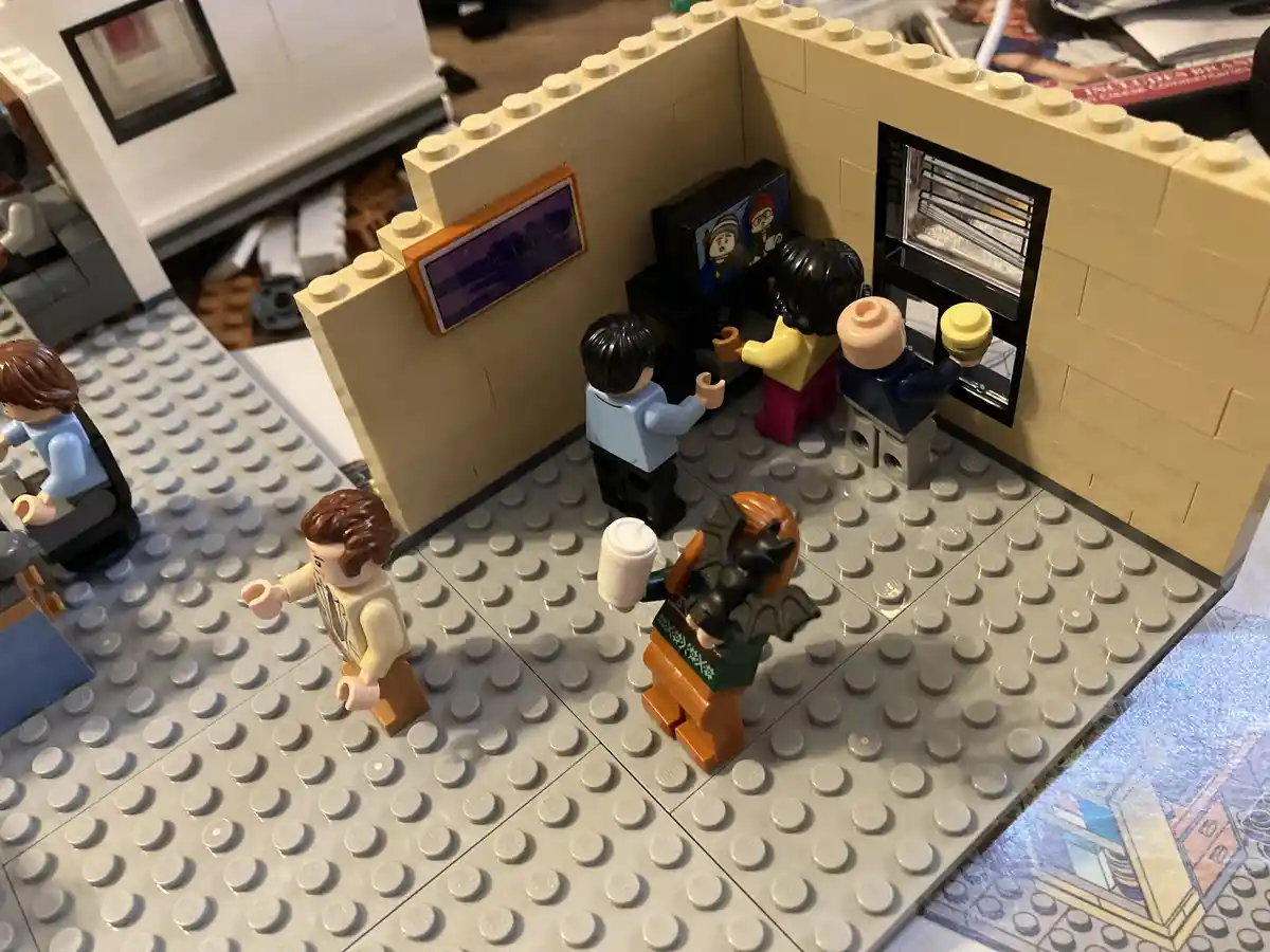 A Lego diorama depicts a living room scene with several Lego minifigures. One figure in a green sweater with bat wings sits…