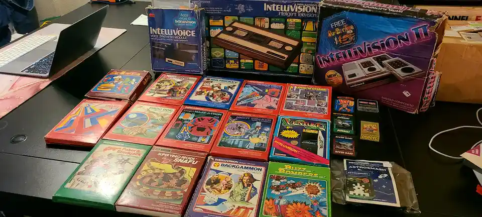 A collection of vintage Intellivision video game consoles and cartridges are displayed on a desk, including boxes for the…