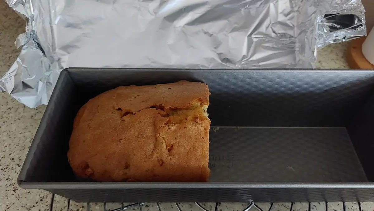 A partially baked loaf cake sits in a metal loaf pan, occupying only about half the pan's space, with the other half empty…