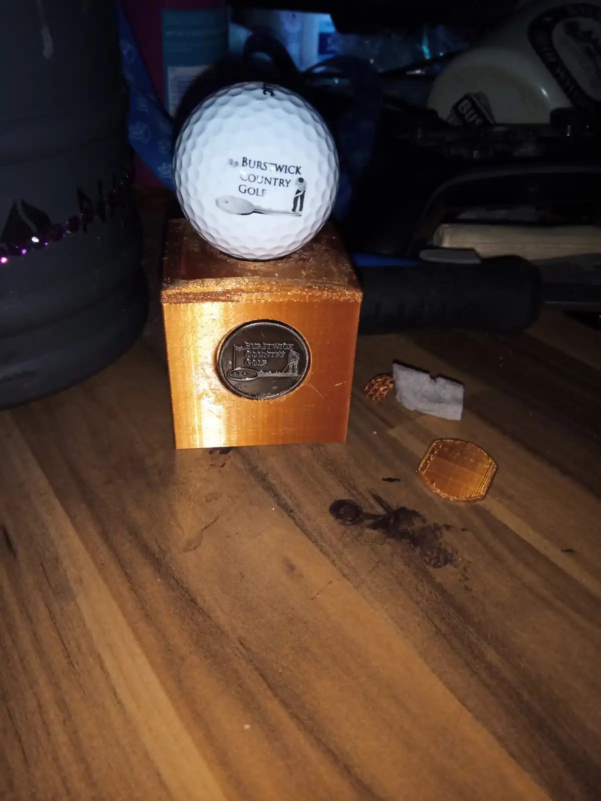 A white golf ball with 'BURSTWICK COUNTRY GOLF' text and a spoon graphic sits atop a small, gold-painted wooden block that…