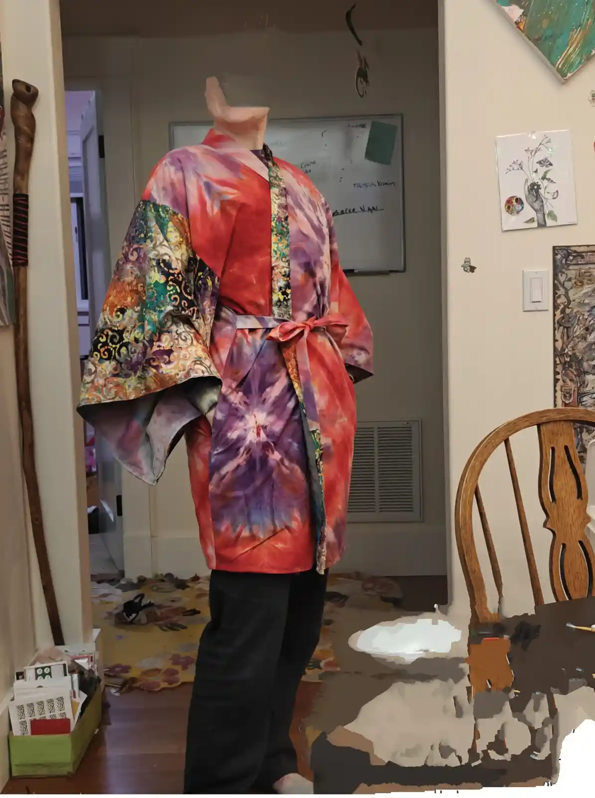 A person stands in a room wearing a brightly colored, patchwork kimono with tie-dye and ornate patterns, paired with black…