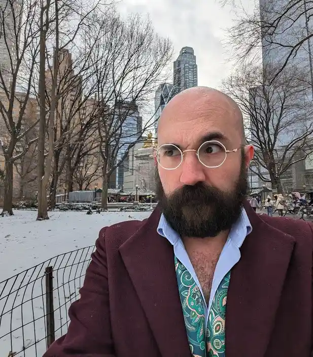 A man with a bald head, thick beard, and round glasses stares sideways with a skeptical expression while wearing a maroon…