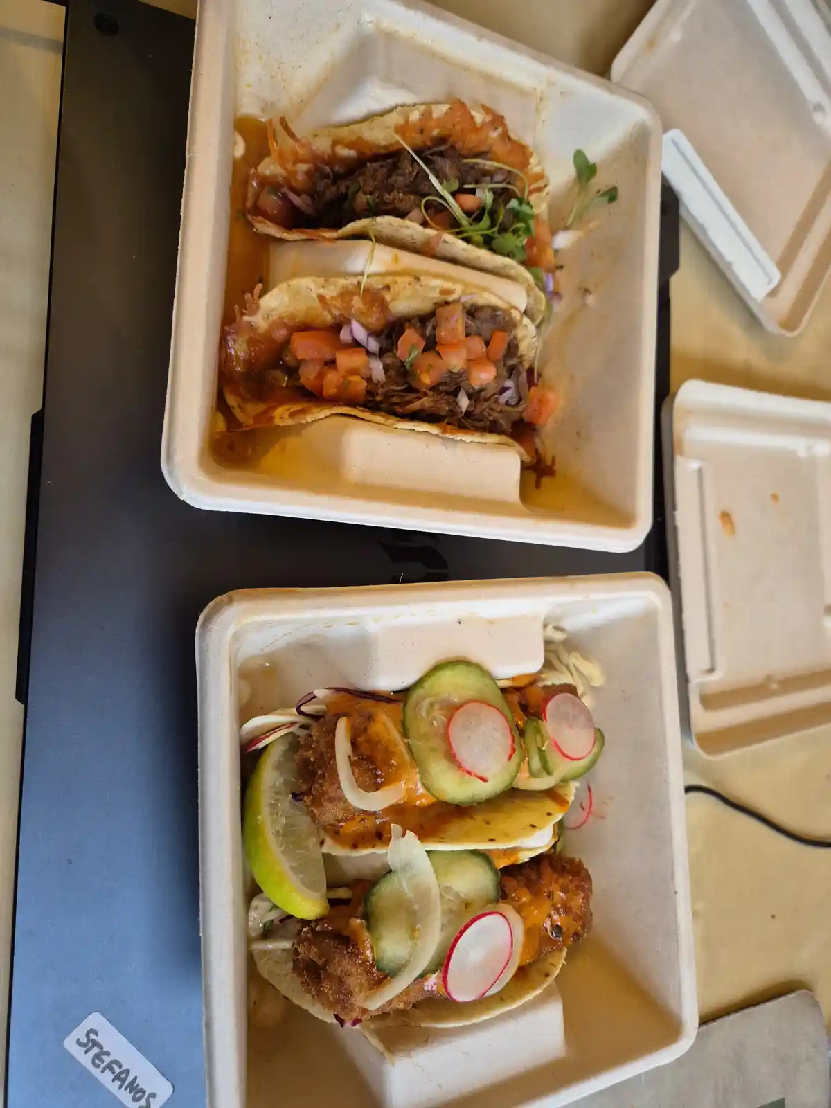 Two takeout containers hold tacos: one with shredded meat and pico de gallo, the other with fried chicken, radishes, and…