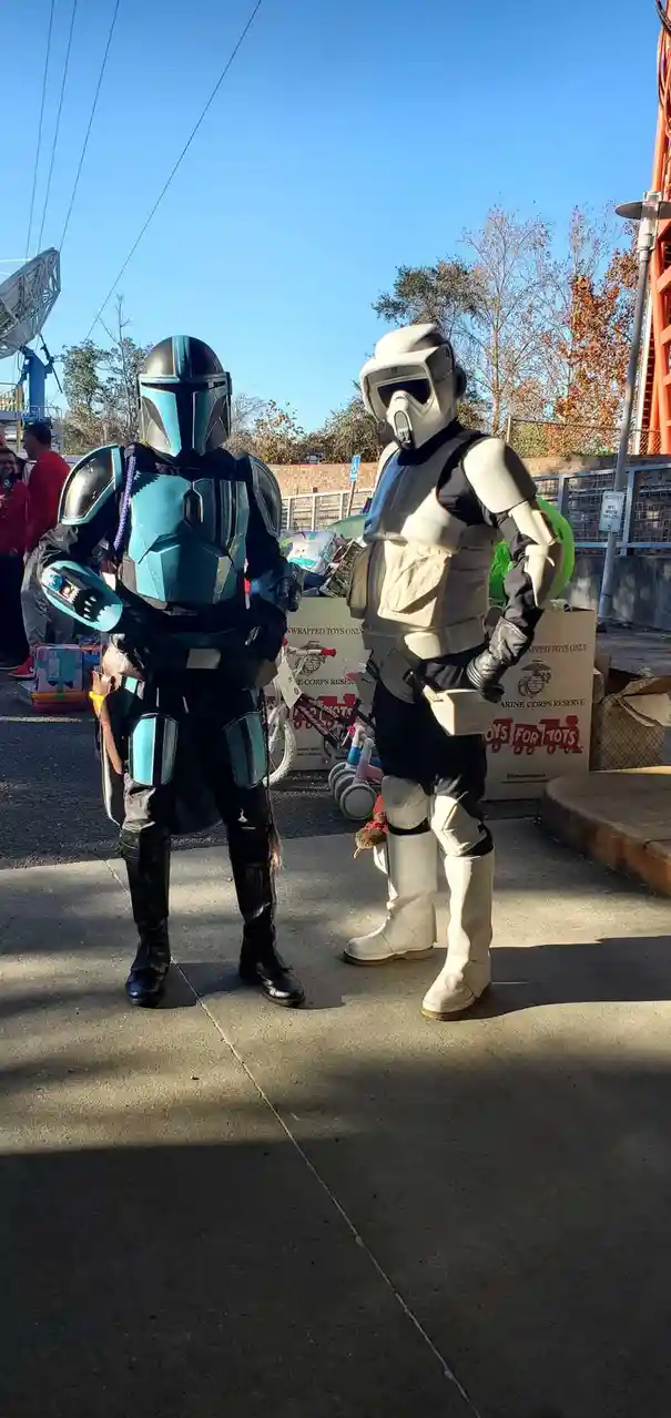 Two people in Star Wars cosplay stand side by side outdoors near a 'Toys for Tots' donation bin. The person on the left…