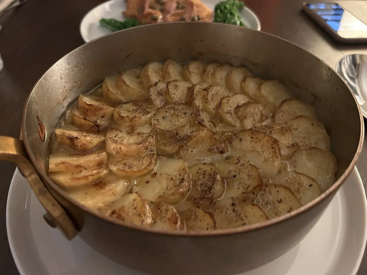 A close-up photo shows a copper-colored pan filled with neatly arranged, sliced, and sautéed potatoes in a creamy sauce…