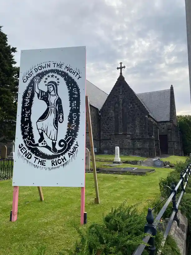 A large, light-blue protest sign stands in a grassy cemetery, featuring a black-and-white illustration of a woman in a robe…