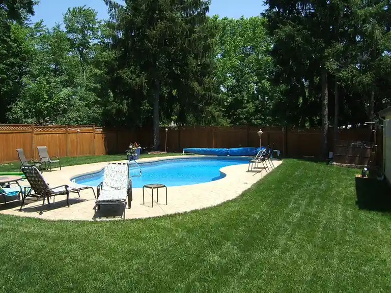 A backyard with a clear blue swimming pool surrounded by a stone patio, lounge chairs, and green grass, with a wooden fence…