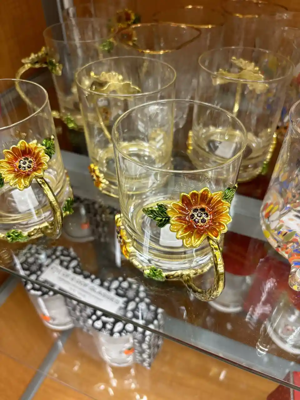 A close-up shot of ornate glass mugs with gold handles shaped like sunflowers, displayed on a glass shelf in a store. The…