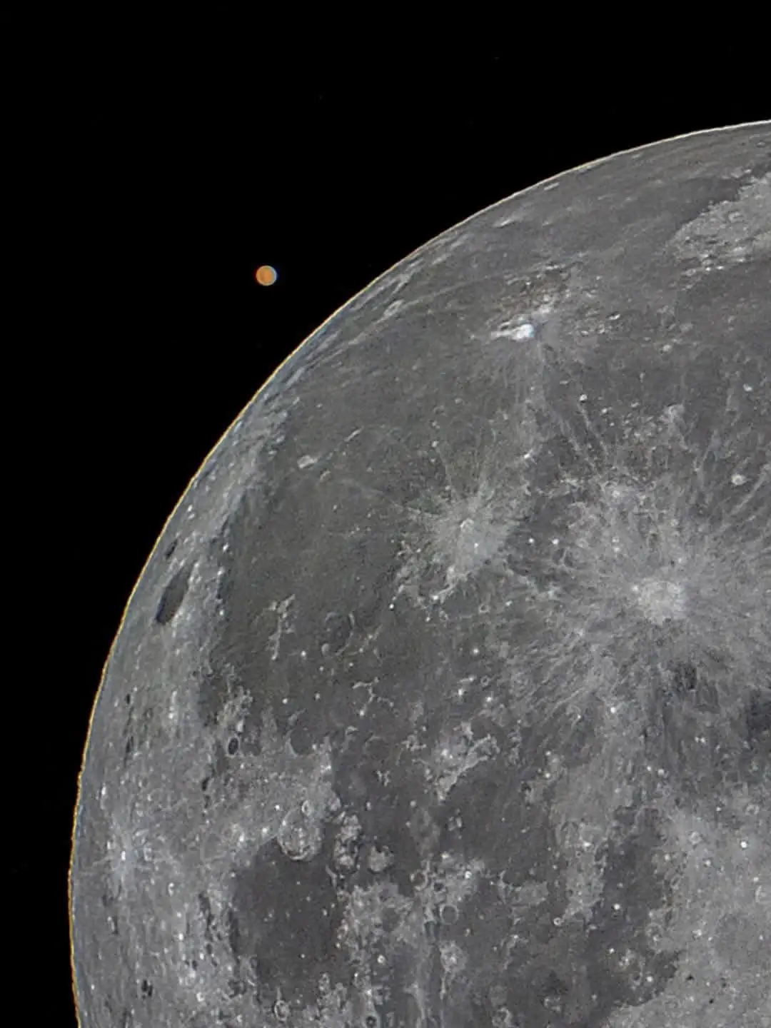 A close-up image of the moon's cratered gray surface against a black sky, with a small orange planet visible near the moon's…