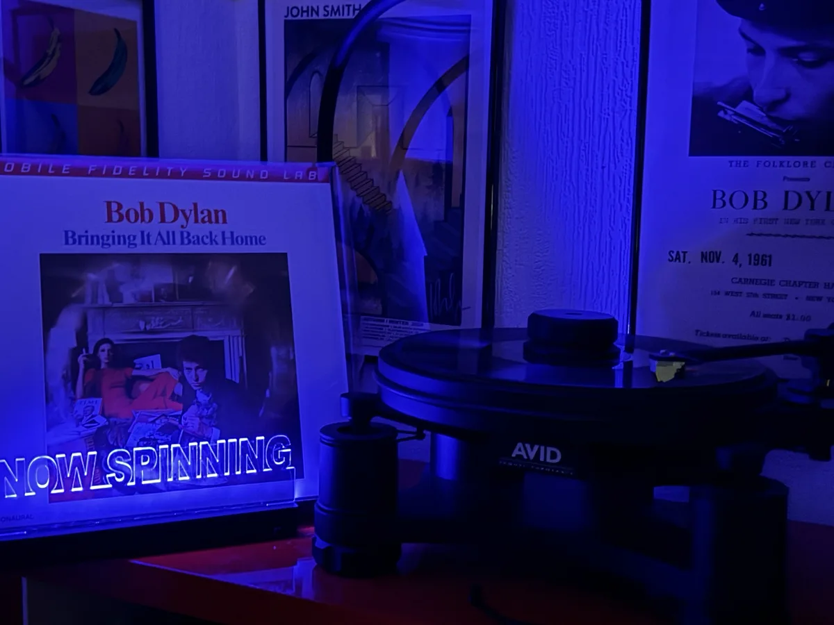 A dimly lit room bathed in blue light features a turntable with a record on it, next to a Bob Dylan album cover for…