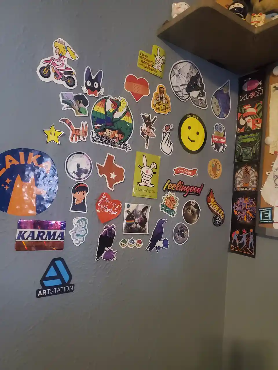 A gray wall is densely covered with dozens of colorful stickers featuring anime characters, pop culture icons, animals, and…