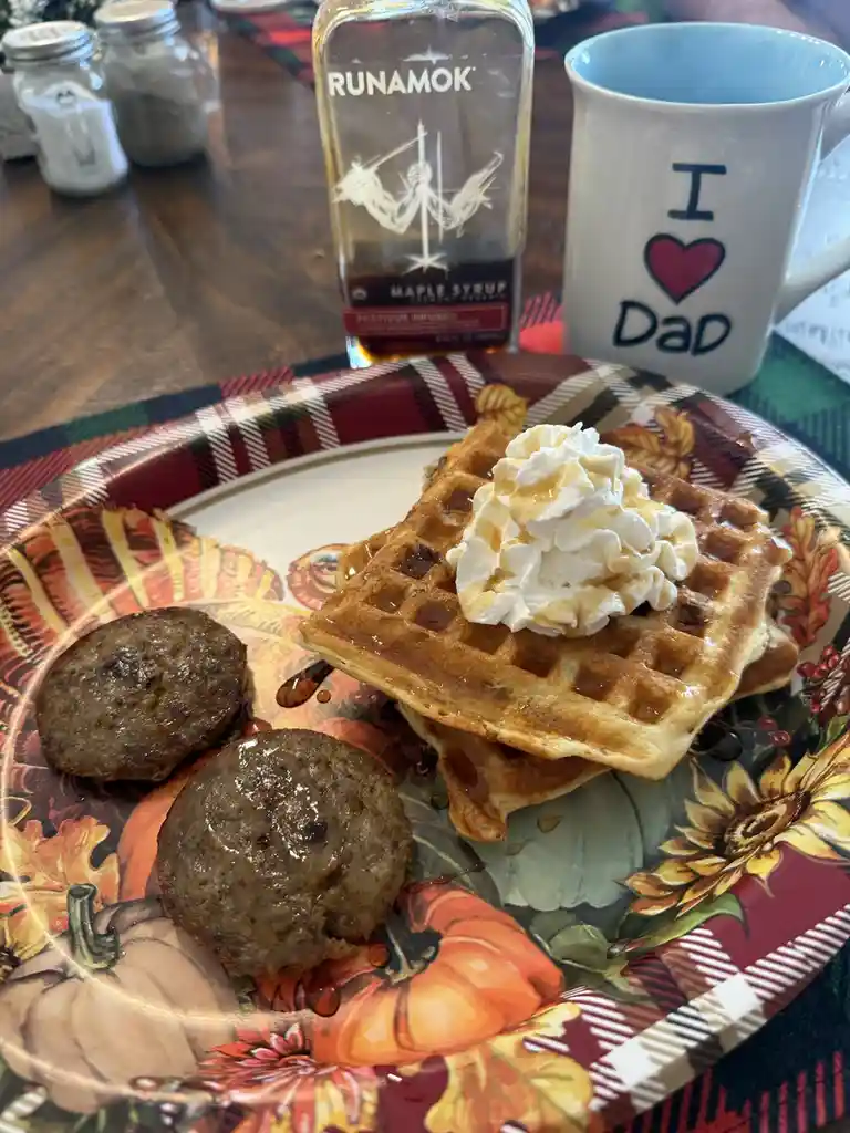 A plate of waffles topped with whipped cream and syrup, accompanied by two sausage patties, sits on a fall-themed plate. In…