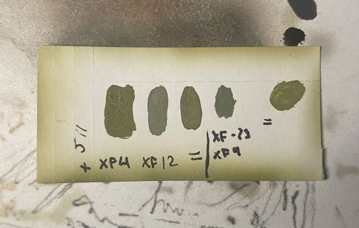 A close-up of a yellowish paper strip with five green paint swatches arranged horizontally. Handwritten text below reads…
