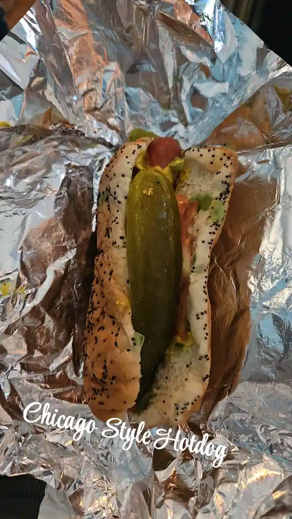 A close-up photo shows a Chicago-style hot dog in a poppy seed bun, nestled in crinkled aluminum foil. The hot dog is topped…