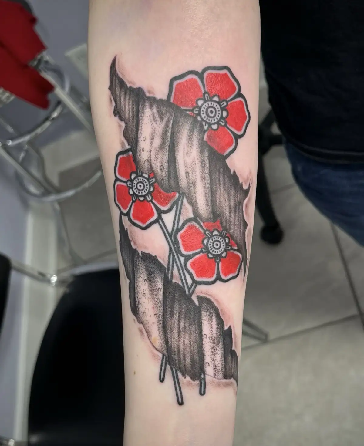 A close-up photo shows a freshly inked tattoo on a person's forearm featuring three bright red poppies with black outlines…