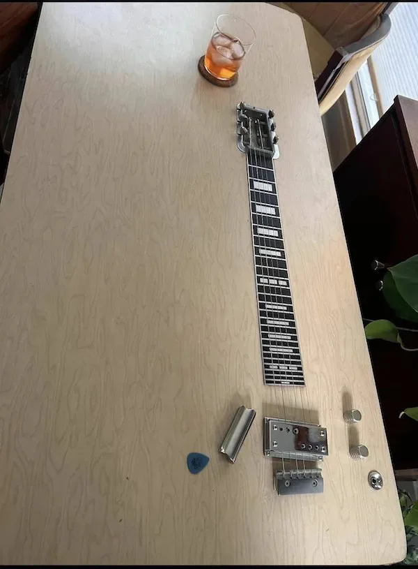 An electric guitar is laid flat on a light wood table, its neck pointing toward a glass of iced tea. A blue guitar pick and…