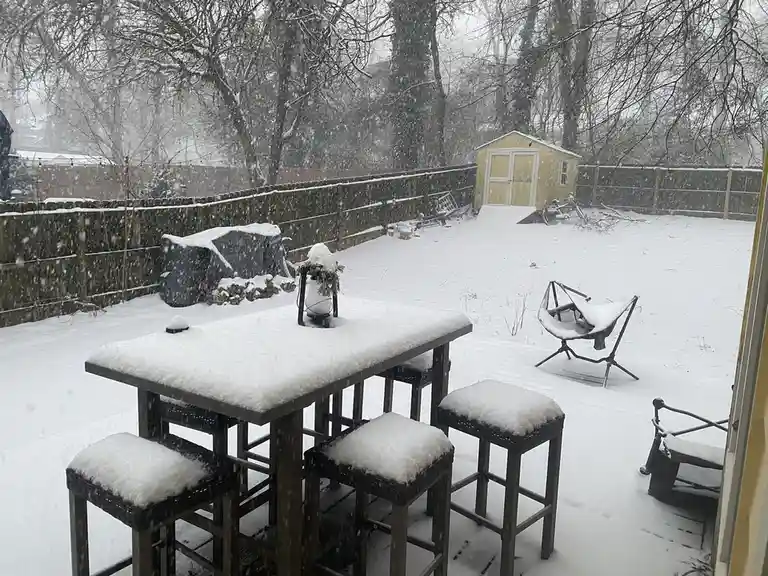 A snowy backyard scene viewed from a window, showing a snow-covered outdoor table and bar stools, a hammock chair, a shed…