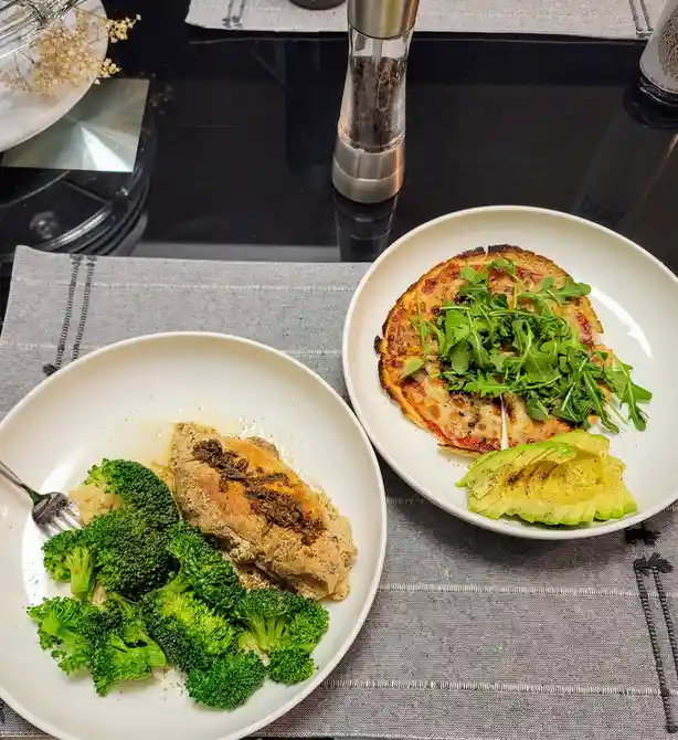 Two white plates sit on a grey placemat on a black countertop. The left plate holds a piece of cooked chicken with broccoli…