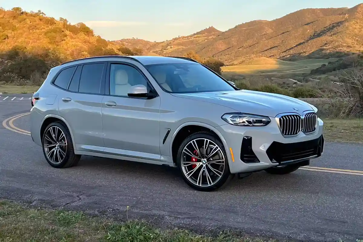 A silver BMW X3 SUV is parked on a paved road with rolling hills and golden-hour lighting in the background. The car…