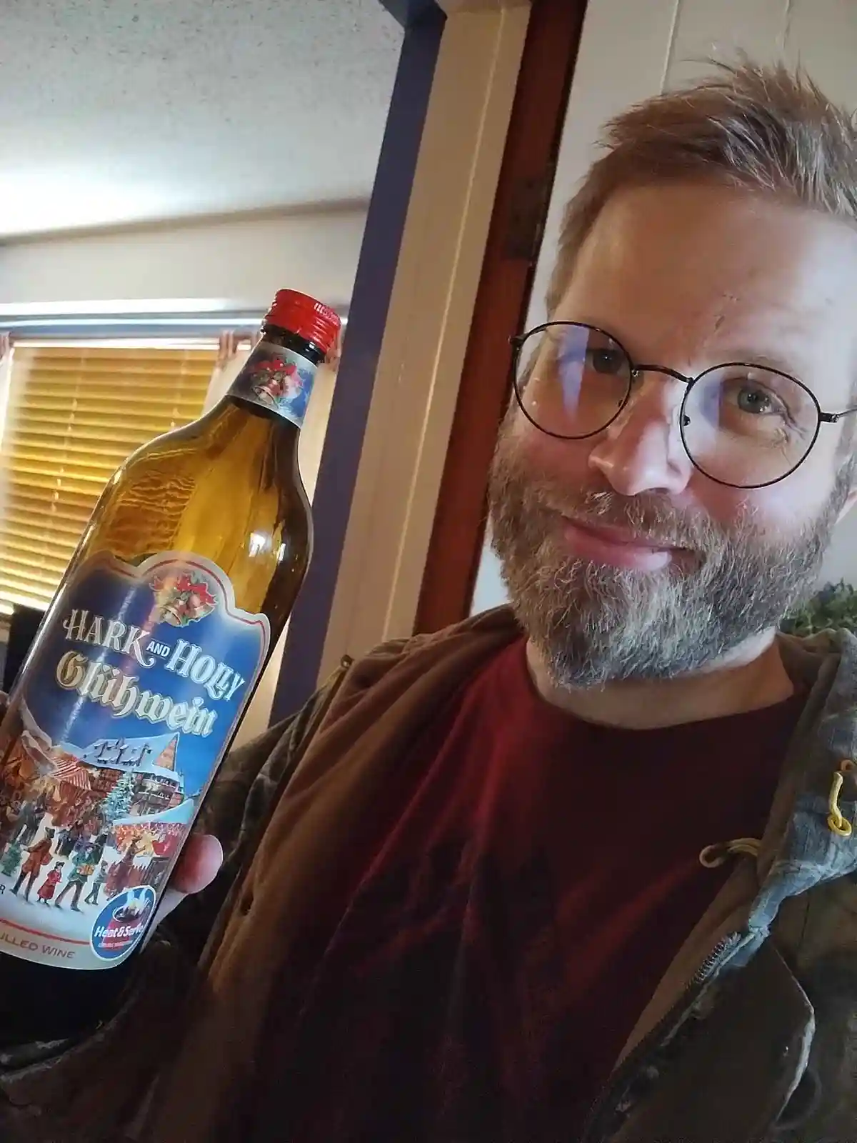 A man with glasses and a beard holds up a bottle of 'Hark and Holy Glühwein' wine, smiling at the camera in a selfie-style…