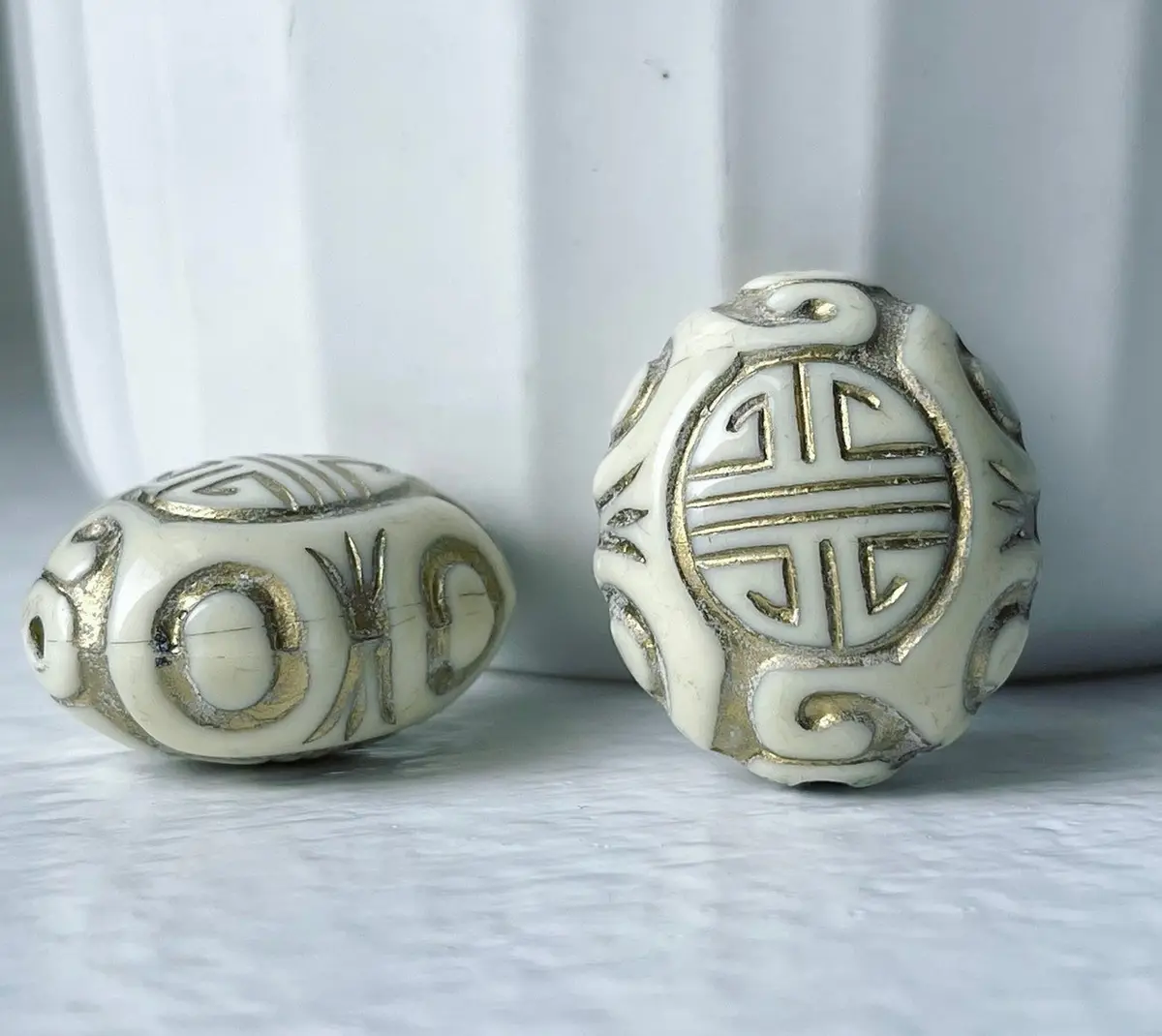 Two ornate, off-white beads with gold filigree patterns sit on a light gray surface; one bead shows a circular geometric…