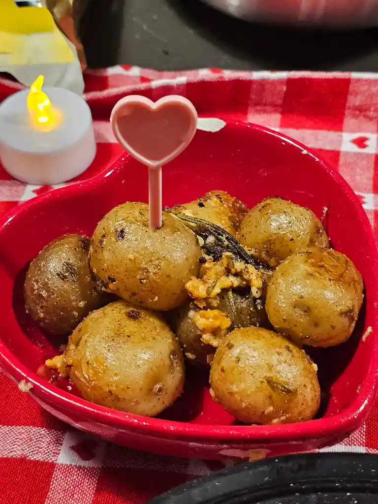 A red heart-shaped bowl filled with roasted baby potatoes sits on a red and white checkered tablecloth, adorned with a pink…