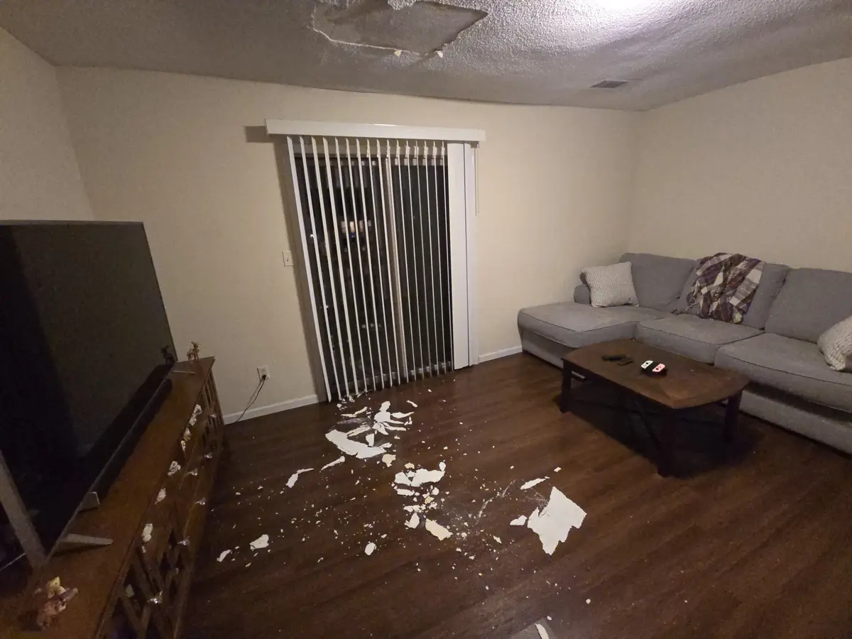 A living room with a large section of ceiling drywall missing and falling debris scattered across the dark wood floor, near…