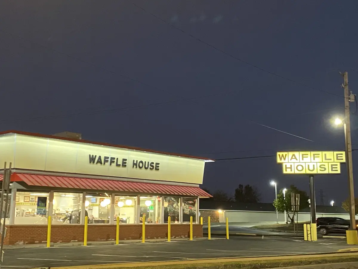 An exterior shot of a Waffle House restaurant at night, showing its illuminated sign and building facade with red awnings…