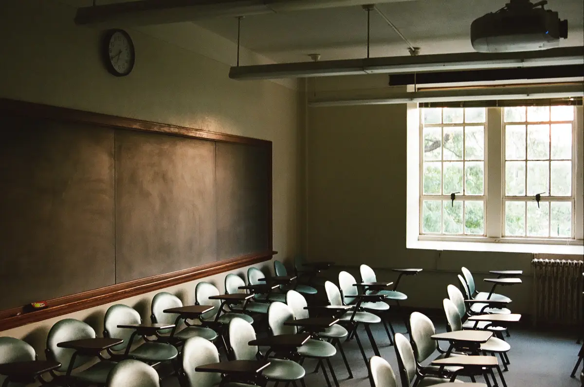 An empty, dimly lit classroom with rows of desks and chairs facing a large blackboard, a clock on the wall, and a projector…