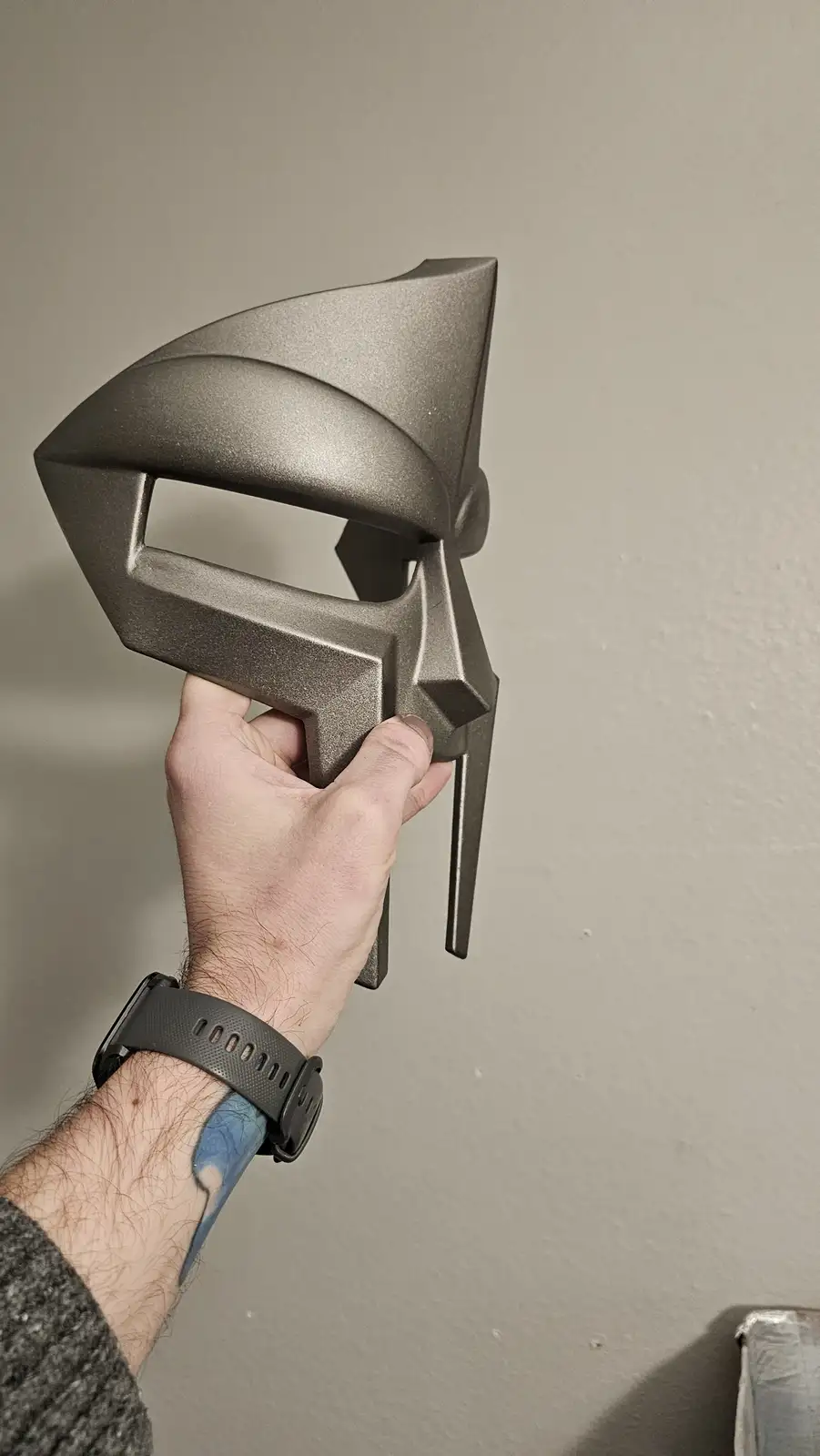A hand holds up a gray, angular, 3d-printed mask resembling a stylized helmet or faceguard against a plain wall. The mask…