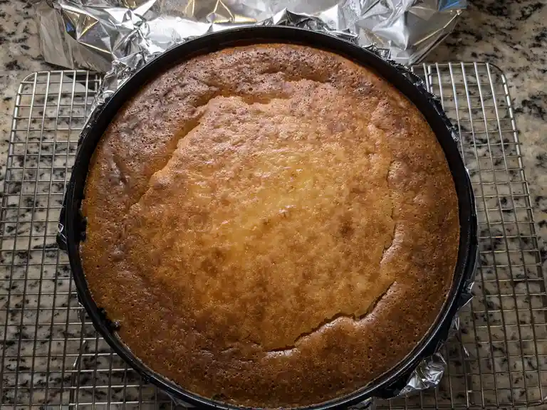 A freshly baked cake in a round metal pan sits on a wire cooling rack over a granite countertop, with aluminum foil…