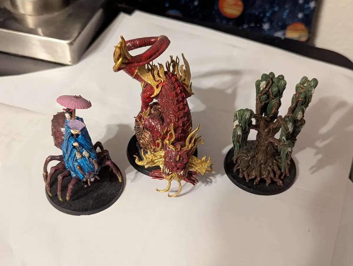 Three painted fantasy miniatures sit on a white surface: a geisha riding a spider, a red and gold dragon, and a gnarled tree…
