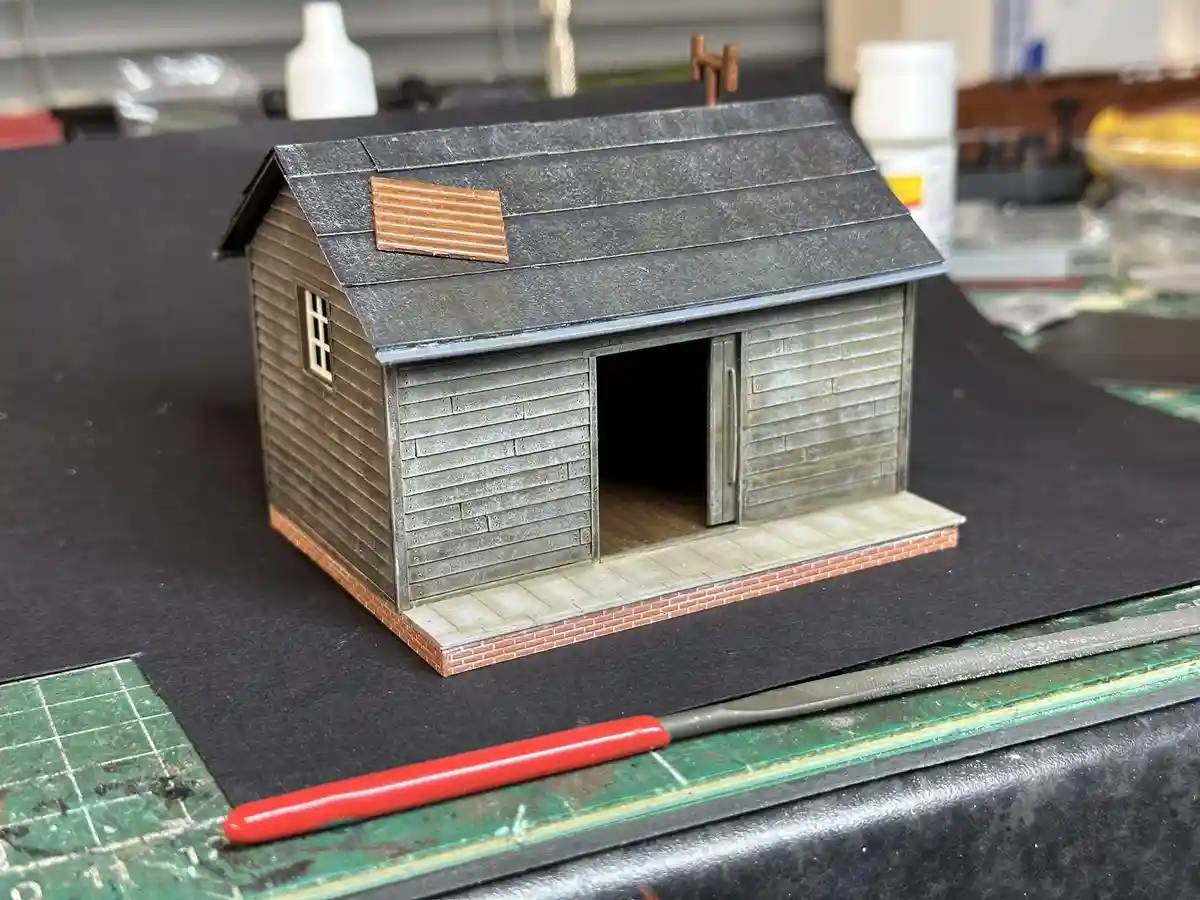 A detailed miniature model of a wooden shed with a gray roof, a small window, and an open door sits on a black mat. A…