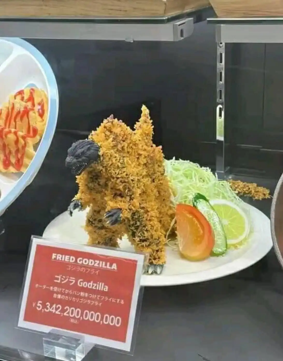 A plate of fried Godzilla-shaped food, covered in breadcrumbs and garnished with vegetables, is displayed with a red sign…