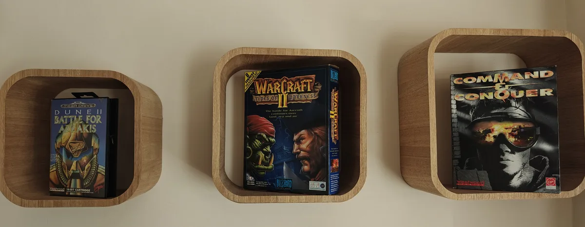 Three wooden wall shelves display classic PC strategy game boxes: Dune II (left), Warcraft II: Tides of Darkness (center)…
