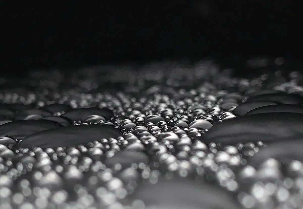 A macro photograph shows countless glistening water droplets on a dark, textured surface, with shallow depth of field…