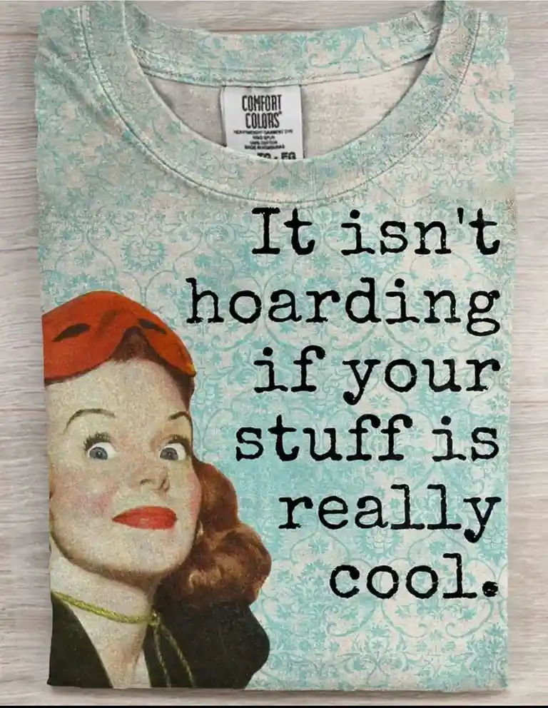 A folded light blue t-shirt with a vintage damask pattern displays the text 'It isn't hoarding if your stuff is really…