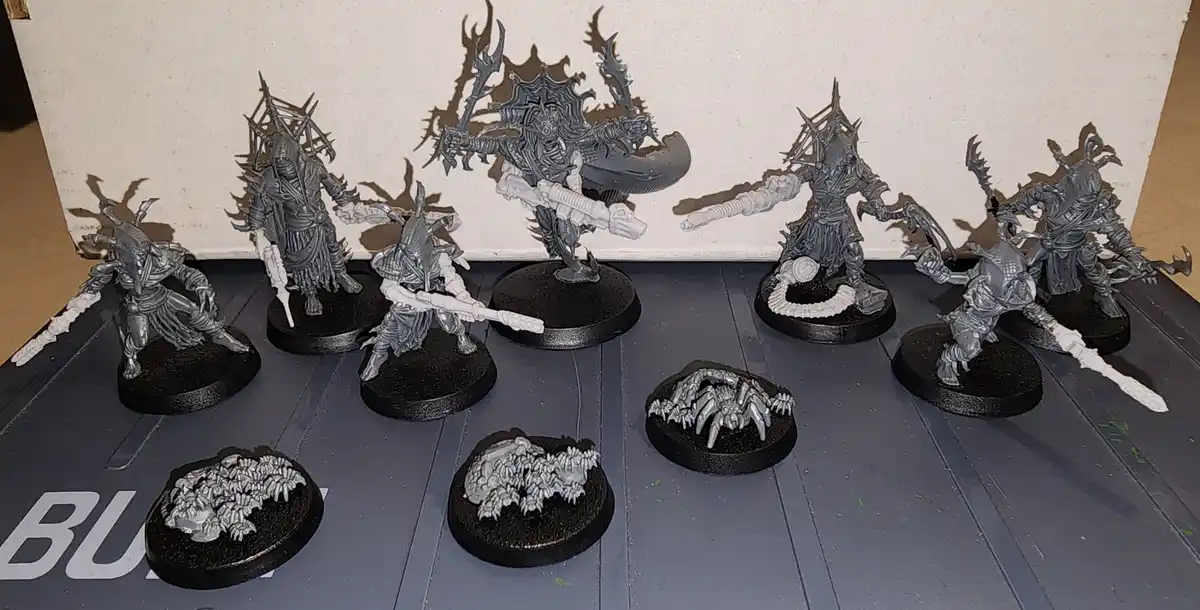 A collection of unpainted, grey miniature figurines of fantasy warriors and monsters on black circular bases, arranged on a…