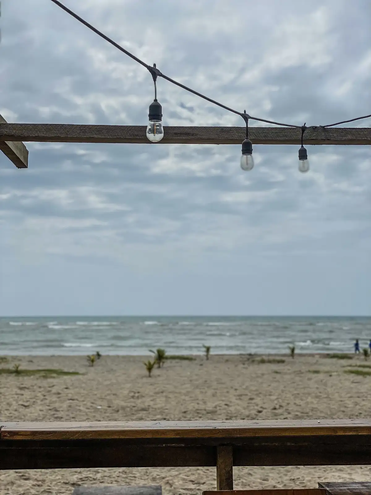 A photo of a beach scene with a wooden railing and string lights hanging above, overlooking the ocean under a cloudy sky…