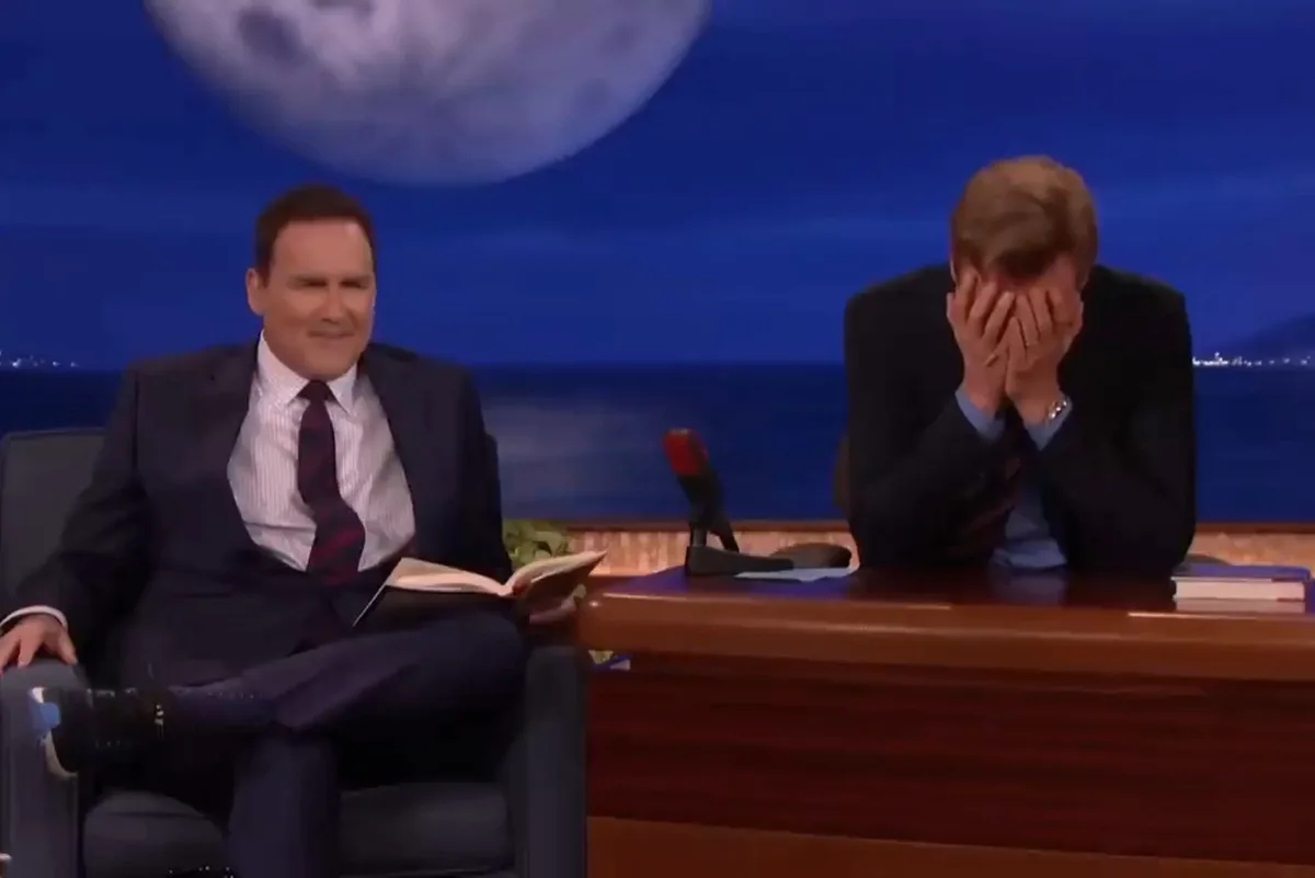 Two men sit on a talk show set; the man on the left smiles smugly while holding a book, and the man on the right covers his…
