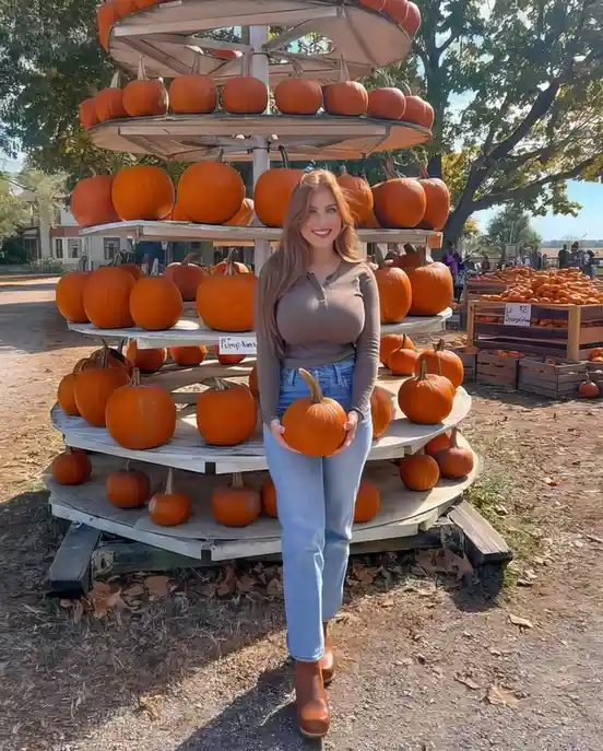 A smiling woman with long blonde hair stands in front of a large, multi-tiered wooden pumpkin display at an outdoor farm…