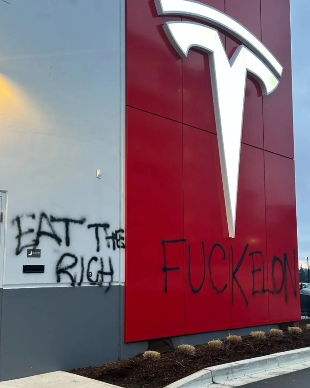 A photo of a Tesla building exterior with a large white Tesla logo on a red wall. Graffiti in black spray paint reads 'EAT…