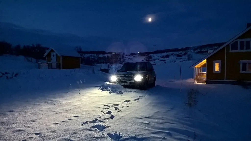 A dark SUV with headlights on is parked in deep snow at night, illuminated by a full moon. Footprints lead toward the…
