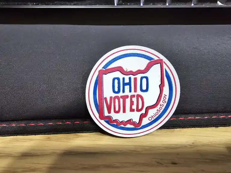 A circular 'OHIO VOTED' sticker with red and blue borders and the state outline of Ohio is placed on a black fabric surface…